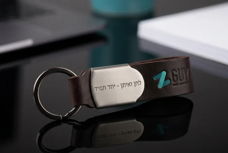 מחזיק מפתחות מעור ומתכת עם חריטה אישית עדינה מונח על שולחן זכוכית שחור.